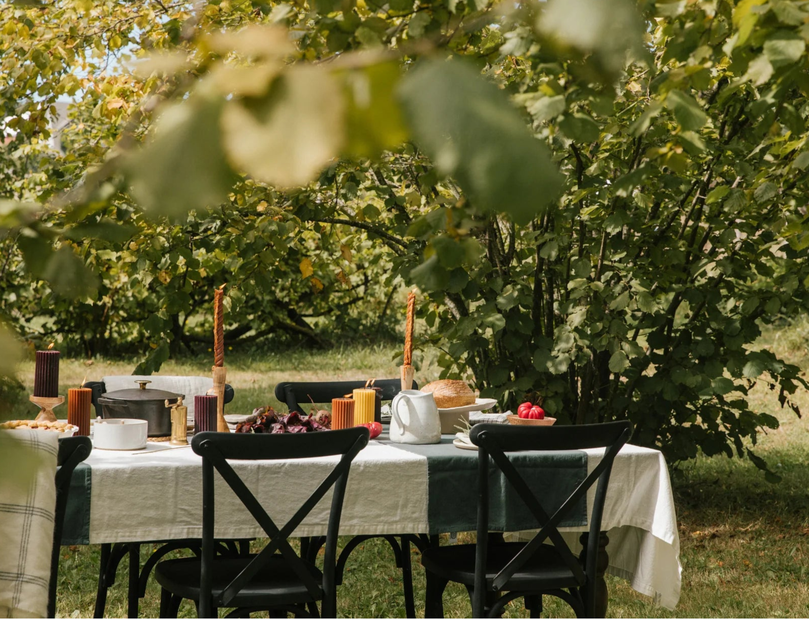A dining table outside in the summer sun set up in a yard. The table is set with food, Crowfoot Collective candles and a table cloth.