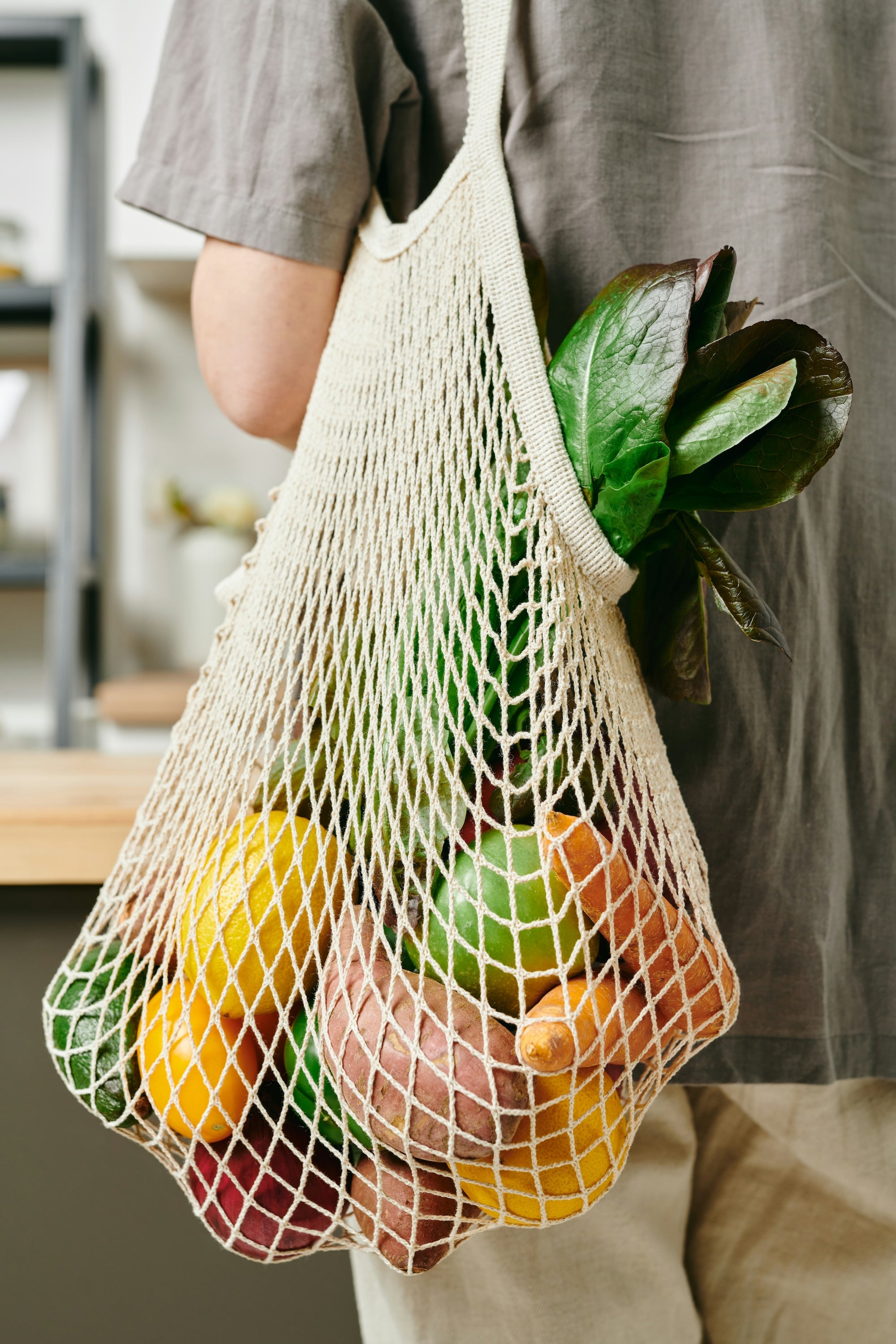 A mesh produce bag is hanging over a persons shoulder while standing in a kitchen. The mesh bag is filled with fresh produce. The linked webpage discuses all of the sustainbility pillars The Good Planet focuses on with the products they offer in store.