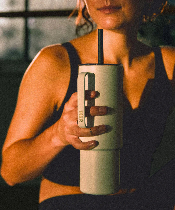 A woman sitting in a room after a workout sipping a beverage from a teal MiiR travel tumbler.
