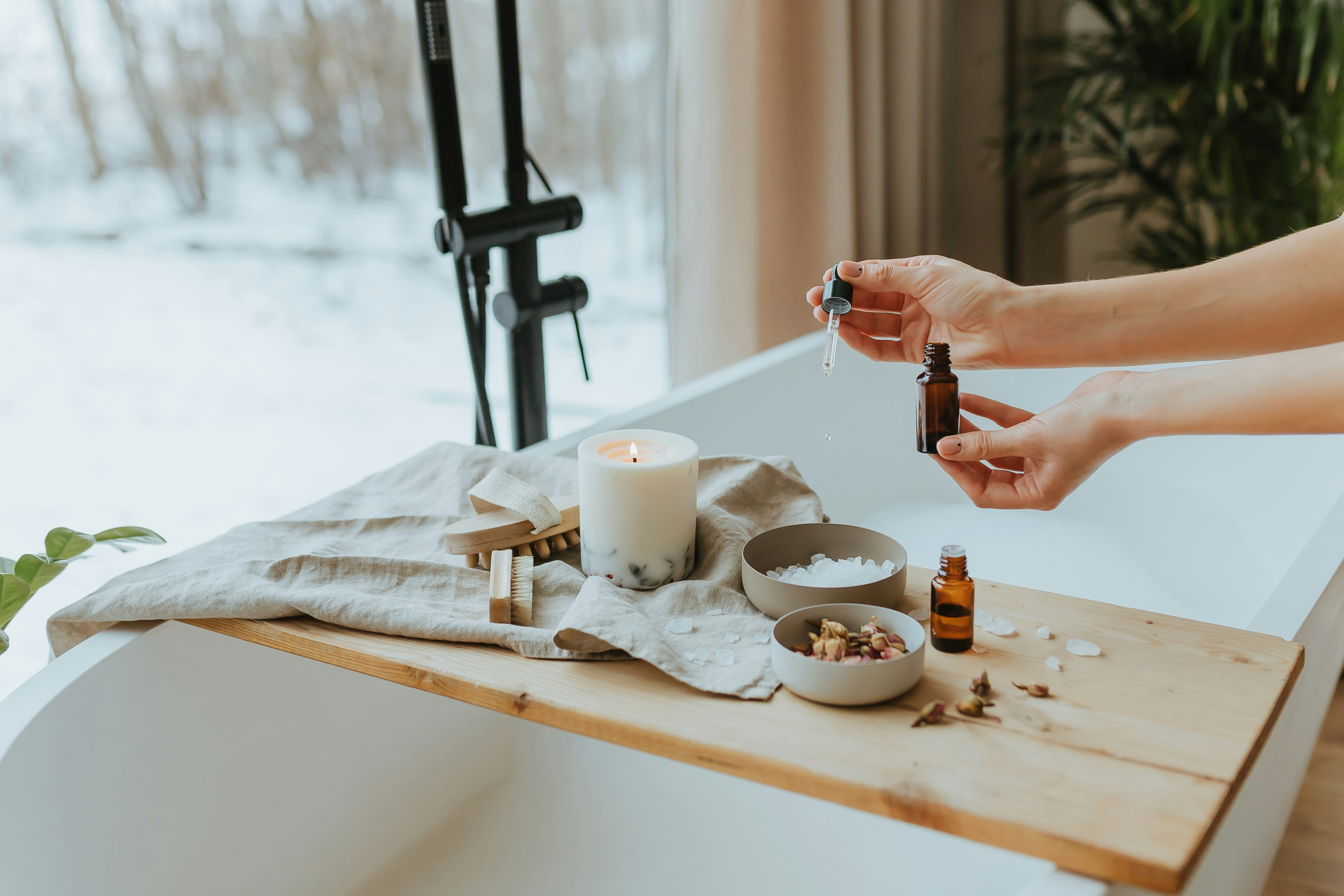 A wooden tray is set across the top of a bathtub. On the tray is an organic linen towel, a foot scrubber, bath massager, a candle, and two bowls with flower petals and bath salts. A person reached over the bowl of bath salts and ads drops of The Good Planets pure essentials oils. 