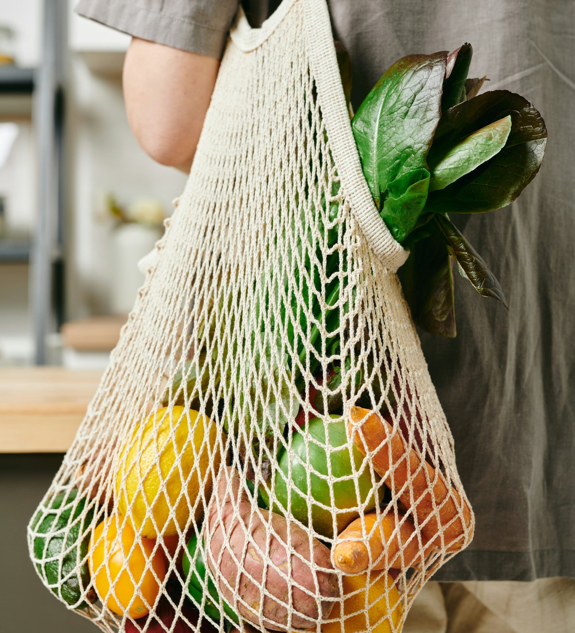 A mesh produce bag is hanging over a persons shoulder while standing in a kitchen. The mesh bag is filled with fresh produce. The linked webpage discuses all of the sustainbility pillars The Good Planet focuses on with the products they offer in store. 