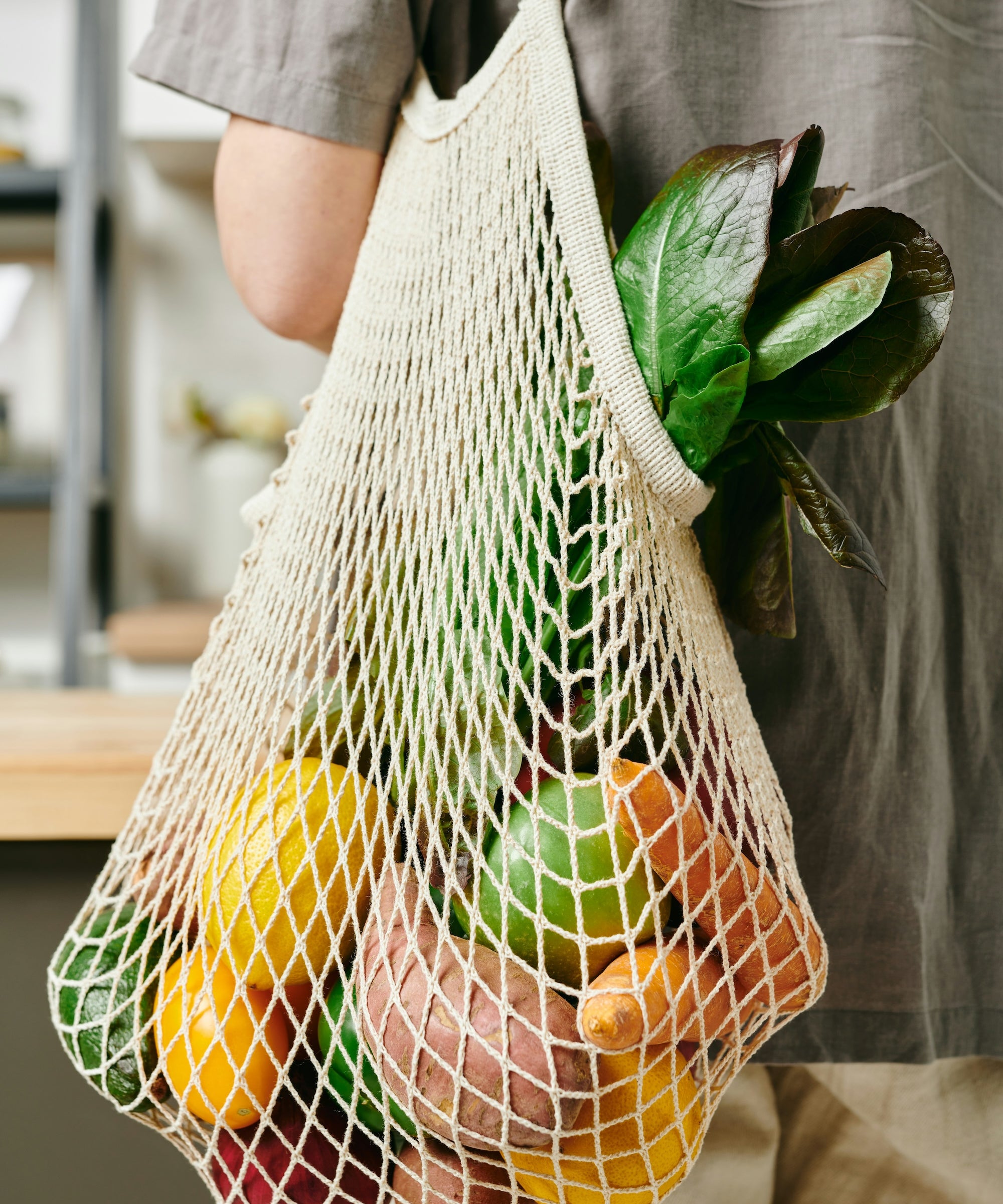A mesh produce bag is hanging over a persons shoulder while standing in a kitchen. The mesh bag is filled with fresh produce. The linked webpage discuses all of the sustainbility pillars The Good Planet focuses on with the products they offer in store. 