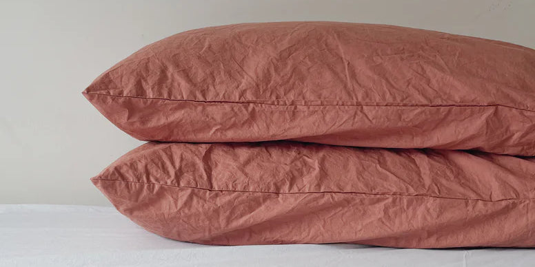 Two pillows are stacked on a bed. They have red colored Good Planet Company pillow cases on them.