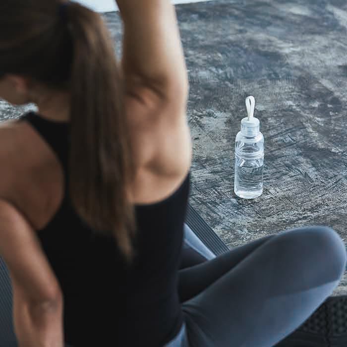 A woman stretches while sitting on a yoga mat with a Kinto water bottle sitting on the ground in front of her. 