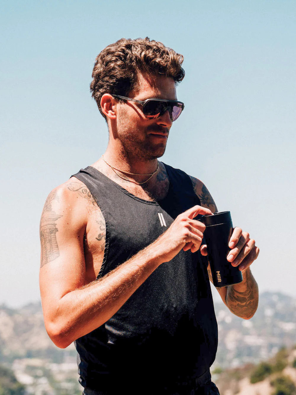 A man standing outside in the sun wearing a tank top and sunglasses opens the lid of his MiiR travel tumbler. 