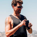 A man standing outside in the sun wearing a tank top and sunglasses opens the lid of his MiiR travel tumbler. 