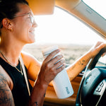 A woman is smiling as she is driving a vehicle while sipping a beverage from her MiiR travel tumbler. 