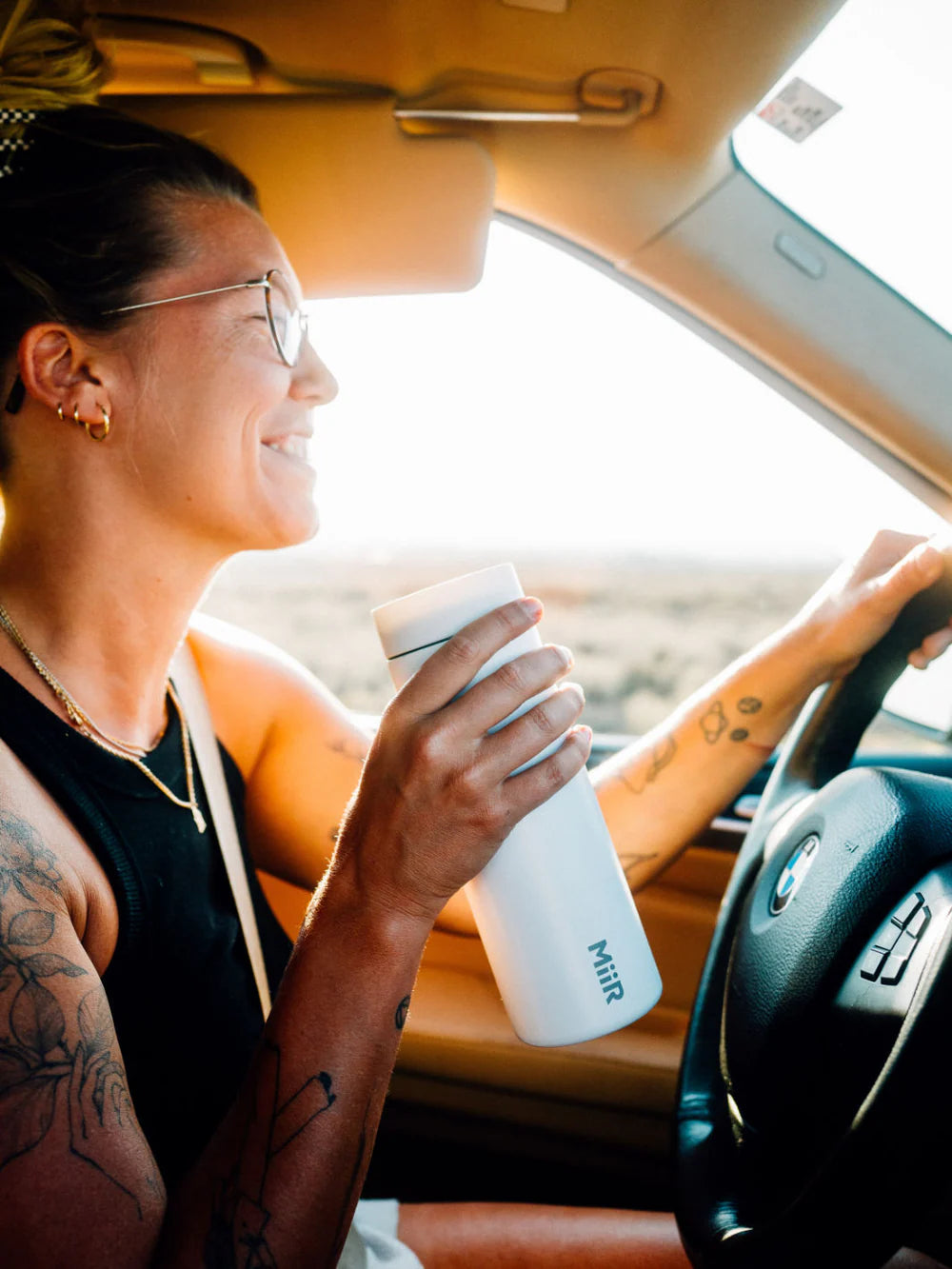 A woman is smiling as she is driving a vehicle while sipping a beverage from her MiiR travel tumbler. 