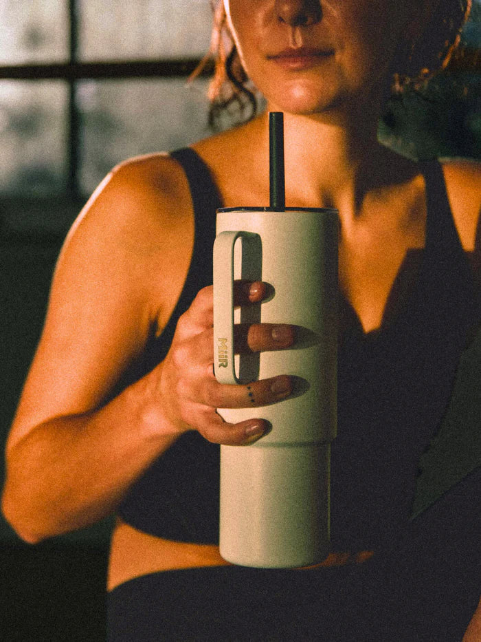 A woman sitting in a room after a workout sipping a beverage from a teal MiiR travel tumbler. 