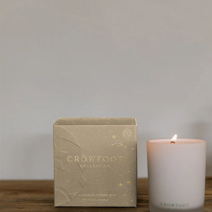 A white glass candle made by Crowfoot Collective sits on a table to the right of the box it was packaged in. 