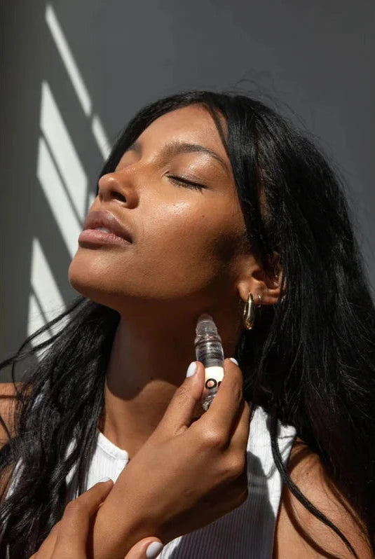 A woman looks up towards the light coming through a window while she applies an OM Organics scented perfume roller onto her next. 